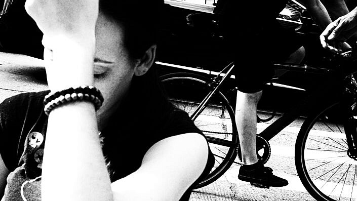 Das Foto zeigt eine junge Frau, die mit geschlossenen Augen und der Hand vor dem Gesicht auf dem Boden am Straßenrad in New York sitzt. Dass Bild ist Teil der Streetphotography Serie von Norbert Smuda aus Dortmund / Deutschland.